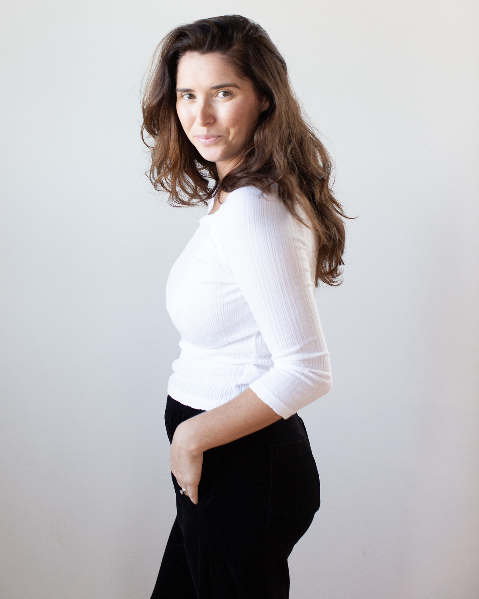 A woman with long brown hair, wearing the Nima L/S Boatneck Top in White by Velvet by Graham & Spencer and black pants, stands in profile against a plain light background, one hand in her pocket and a slight smile on her face.