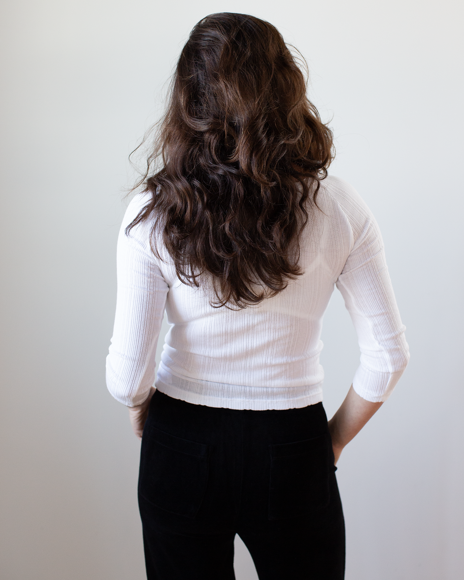 A person with long, wavy brown hair stands facing away from the camera, wearing the Velvet by Graham & Spencer Nima L/S Boatneck Top in White and black pants against a plain light background.