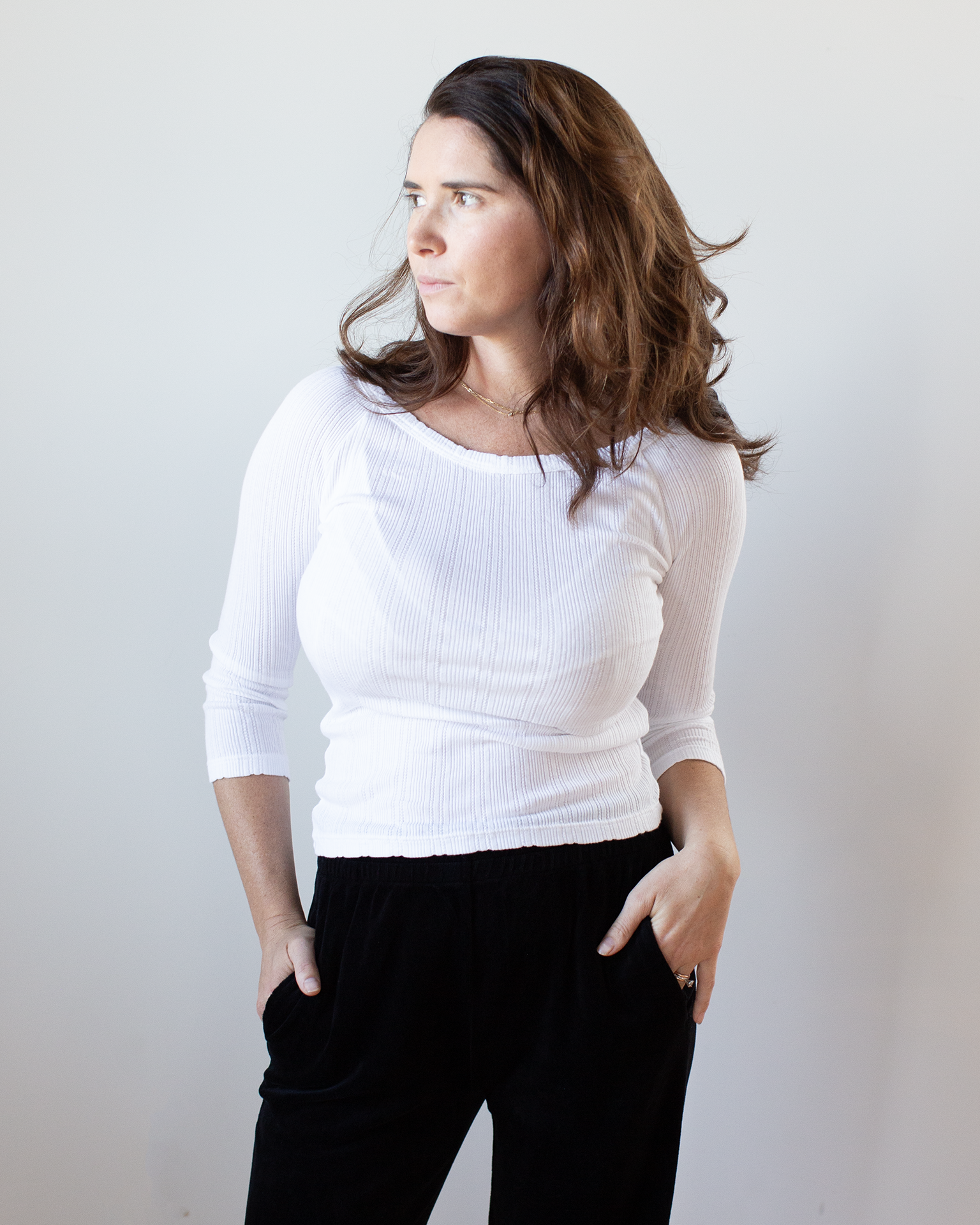 A woman with wavy brown hair wears the Velvet by Graham & Spencer Nima L/S Boatneck Top in White and black pants, standing against a plain light background, looking left with her hands in her pockets.