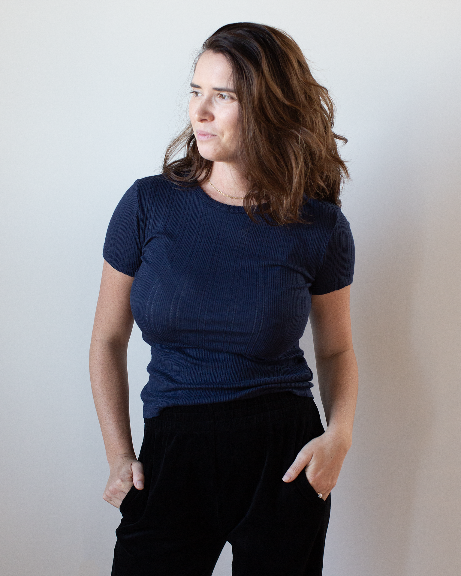 A woman with wavy brown hair wears the Velvet by Graham & Spencer Enati S/S Crew Neck Top in Navy and black pants, standing against a plain light background with her hands in her pockets, looking slightly left and smiling softly.