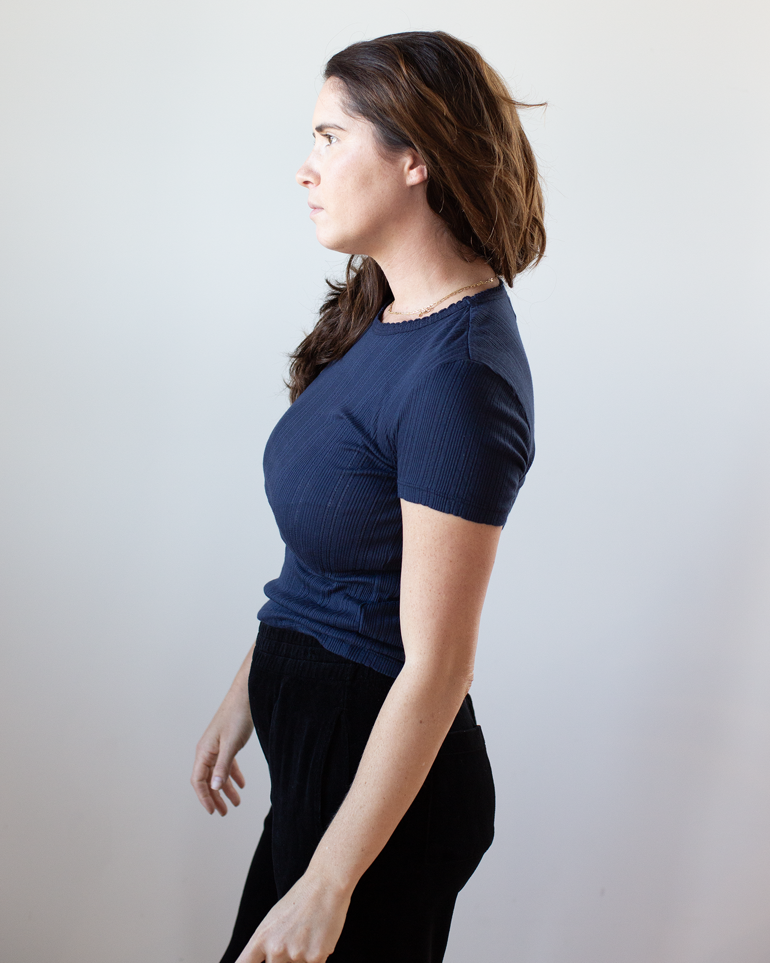 A woman with long brown hair stands in profile against a light background, wearing the Enati S/S Crew Neck Top in Navy and black pants from Velvet by Graham & Spencer.