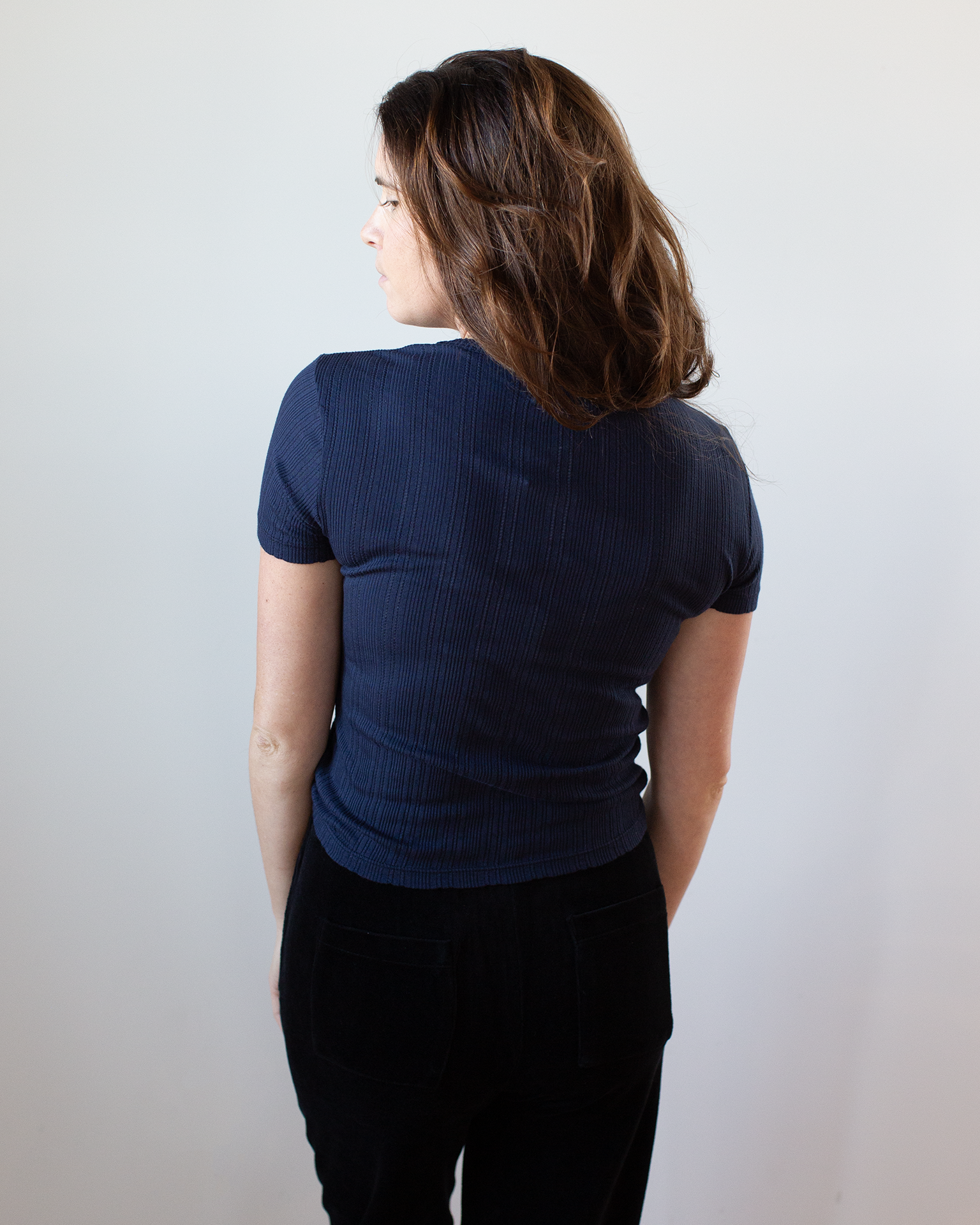 A woman with medium-length brown hair faces away from the camera, wearing a Velvet by Graham & Spencer Enati S/S Crew Neck Top in Navy and black pants against a plain light background.