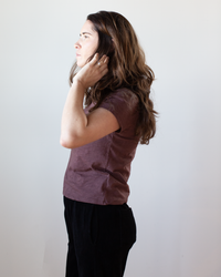 A woman with long brown hair stands in profile against a white background, wearing the Velvet by Graham & Spencer Marie S/S Crop Top in Henna and black pants, with one hand touching her ear.