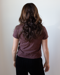 A person with long, wavy brown hair is seen from behind, wearing the Velvet by Graham & Spencer Marie S/S Crop Top in Henna and black pants against a plain light background.