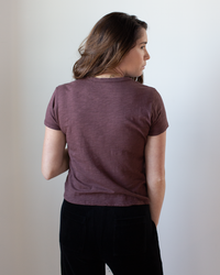 A woman with long brown hair stands facing away, wearing the Velvet by Graham & Spencer Marie S/S Crop Top in Henna and black pants, her right hand in her pocket against a plain white background.