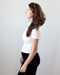 A woman with long brown hair stands in profile against a plain light background, wearing the Enati S/S Crew Neck Top in Ecru by Velvet by Graham & Spencer and black pants, looking ahead with a neutral expression.