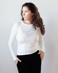 A woman with long brown hair stands against a light background, wearing the Mauri L/S Crew Neck Top in Ecru by Velvet by Graham & Spencer and black pants, looking to her left with her hands in her pockets.