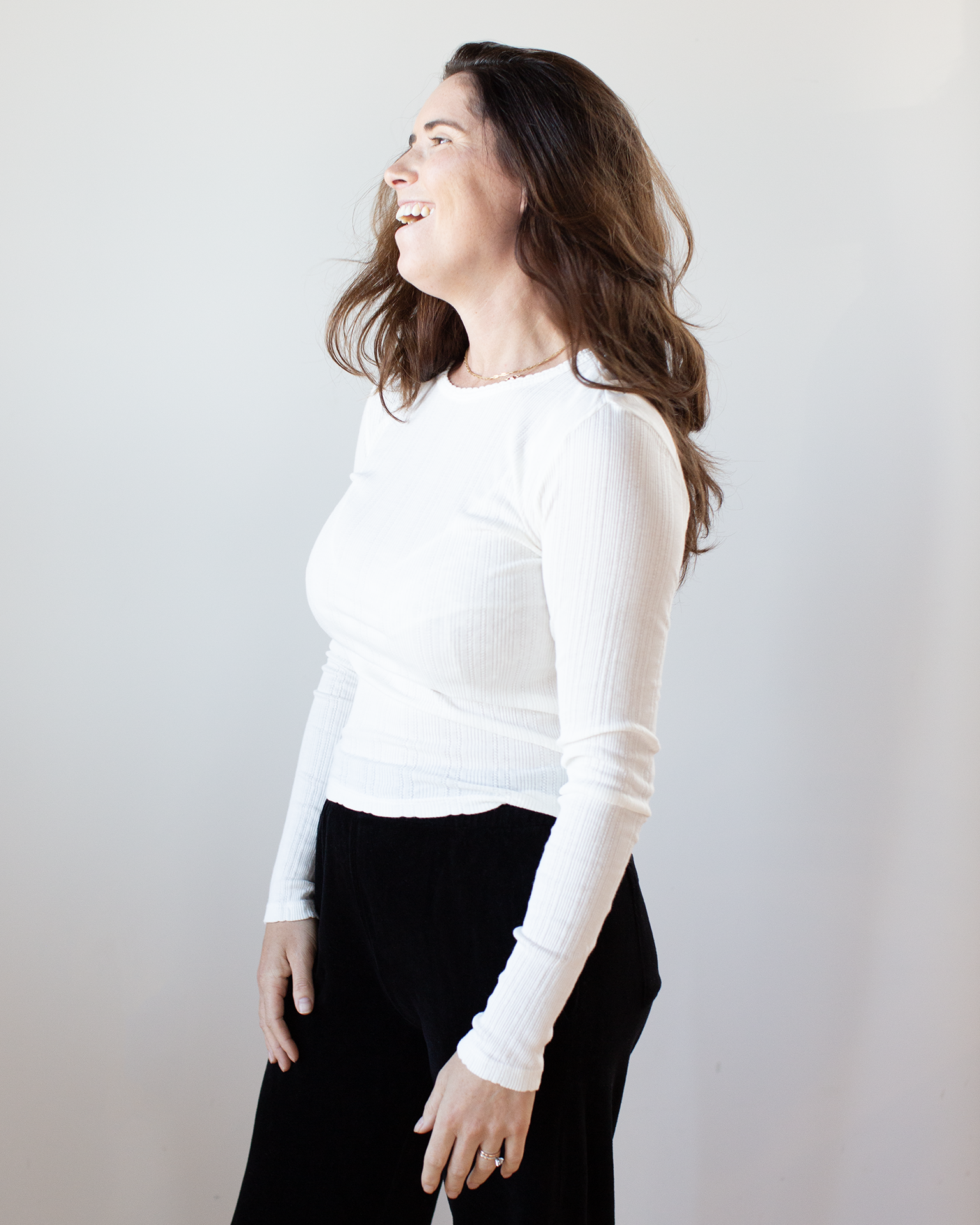 A woman with long brown hair wears the Mauri L/S Crew Neck Top in Ecru by Velvet by Graham & Spencer and black pants, smiling as she looks to her left against a plain light background.