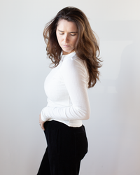 A woman with long brown hair wears the Mauri L/S Crew Neck Top in Ecru by Velvet by Graham & Spencer and slim black pants, standing against a plain light background and looking down with a calm expression.