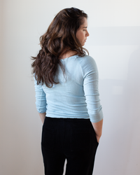 A woman with long brown hair wears the Velvet by Graham & Spencer Nima L/S Boatneck Top in Ceramic and black pants, standing with her back to the camera, hands in her pockets, against a plain white background.