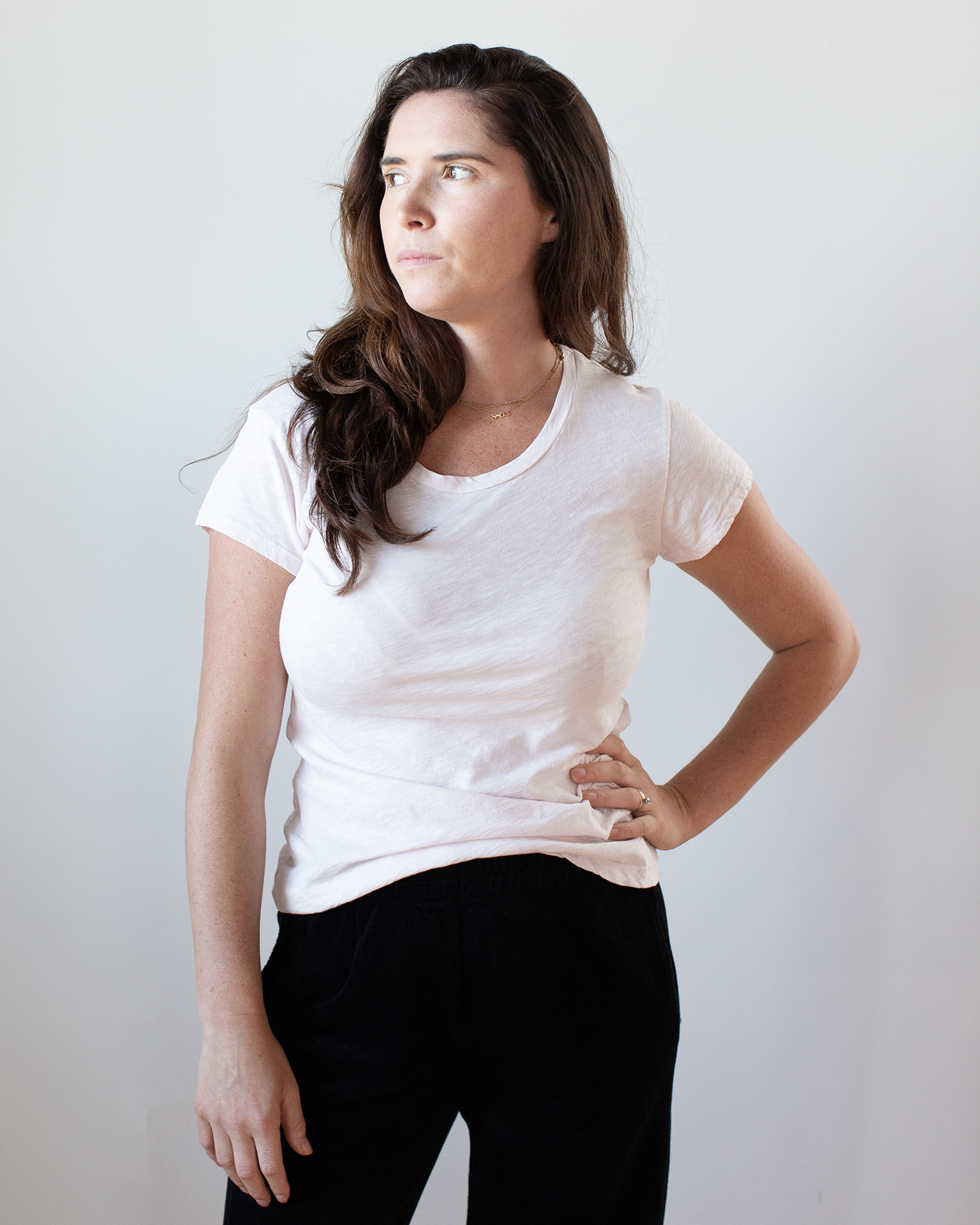 A woman with long brown hair wears the Odelia Scoop Neck Tee in Puff by Velvet by Graham & Spencer and black pants, standing against a plain light background, looking to her left with a neutral expression and one hand on her hip.