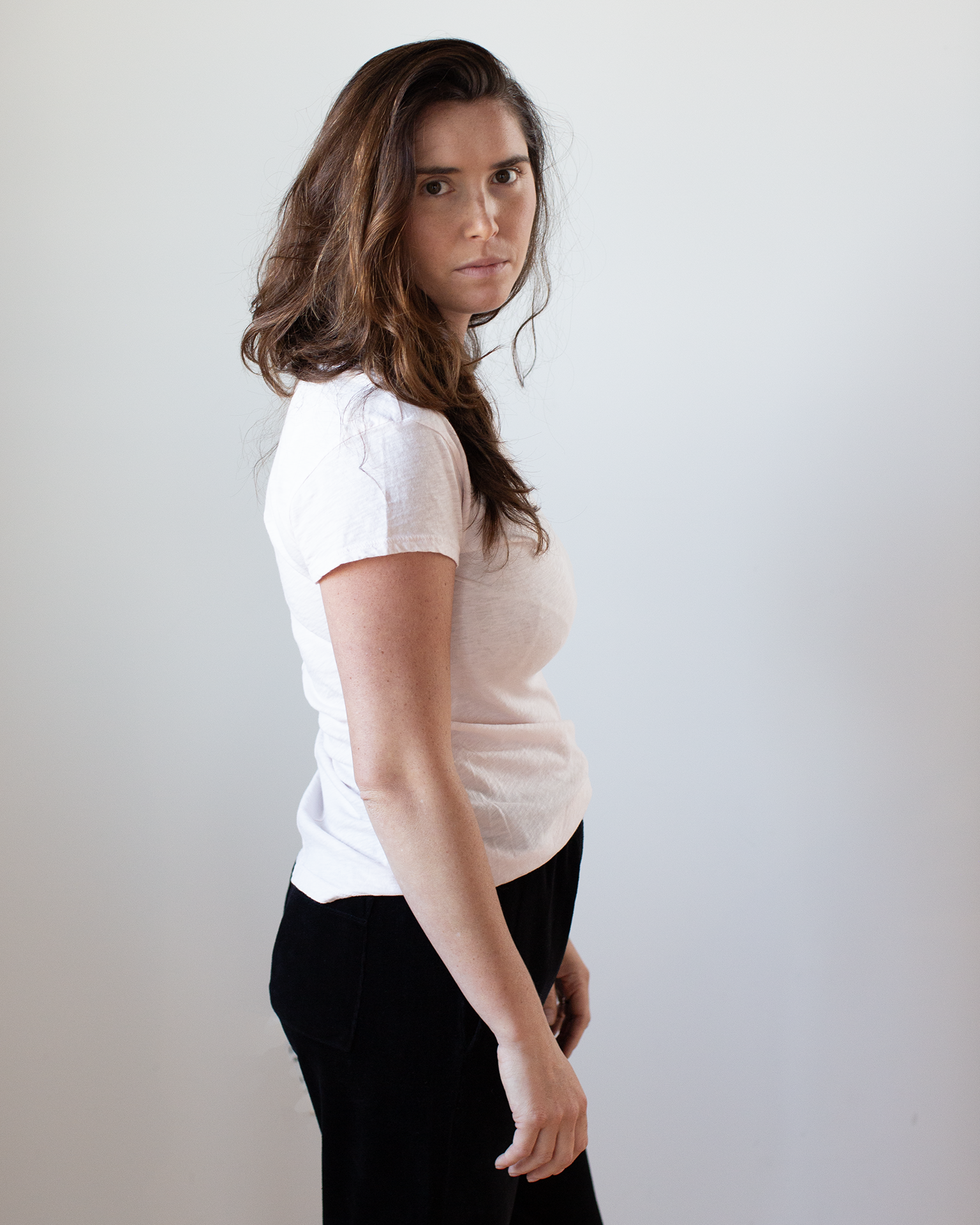 A woman with long brown hair stands sideways, facing the camera with a neutral look. She wears the Velvet by Graham & Spencer Odelia Scoop Neck Tee in Puff and black pants, set against a plain light background.