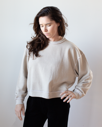 A person with long brown hair wears the NSF Kenya Oversize FT Crew Sweatshirt in Faded Taupe and black pants, standing against a plain, light background and looking down with a neutral expression.