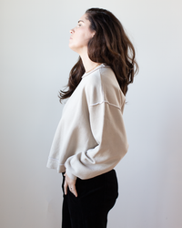 A woman with long brown hair stands in profile against a plain light background, looking slightly upward. She wears the NSF Kenya Oversize FT Crew Sweatshirt in Faded Taupe and black pants.