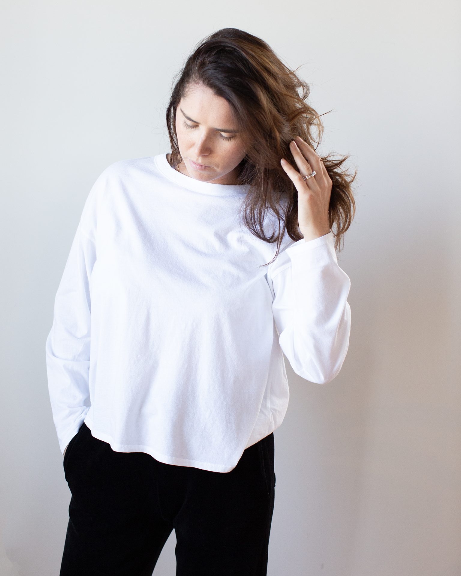 A woman with long brown hair wears the NSF Loffler Boxy Fine Jersey Tee in White and black pants, standing against a plain white background, looking down and touching her hair with one hand.