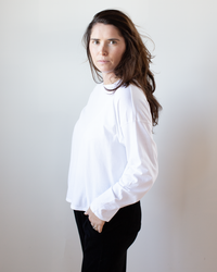 A woman with long brown hair wears the NSF Loffler Boxy Fine Jersey Tee in White and black pants, standing against a light background and looking directly at the camera with a neutral expression.