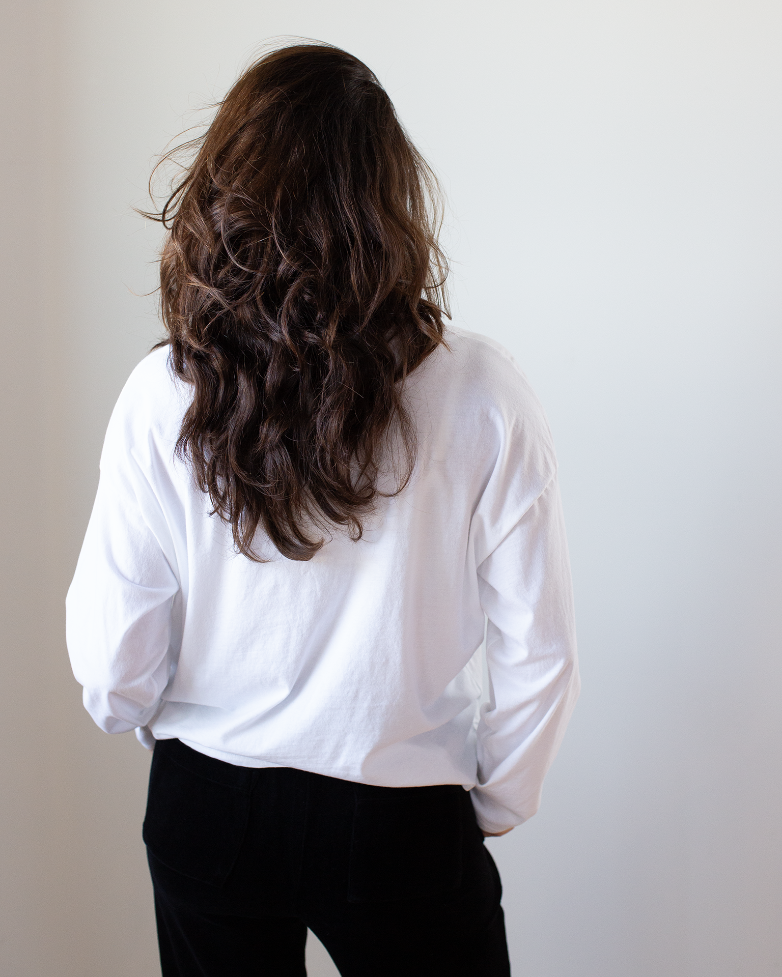A person with long, wavy brown hair stands with their back to the camera, wearing the NSF Loffler Boxy Fine Jersey Tee in White and black pants against a plain light background.