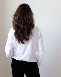 A person with long, wavy brown hair stands with their back to the camera, wearing the NSF Loffler Boxy Fine Jersey Tee in White and black pants against a plain light background.