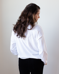 A person with long brown wavy hair, wearing the NSF Loffler Boxy Fine Jersey Tee in White and black pants, stands with their back to the camera, looking slightly to the side against a plain light background.