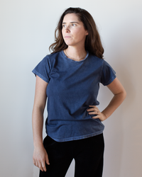 A woman with long brown hair, wearing a Free City Canyonkid Circa1972 Supervintage Tee in Moonshrooms Powder and black pants, stands against a plain light background, looking slightly left with a neutral expression.