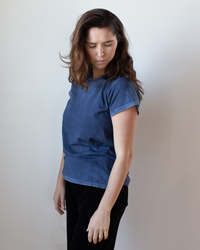 A woman with long brown hair wears the Free City Canyonkid Circa1972 Supervintage Tee in Moonshrooms Powder and black pants, standing against a plain light background, looking down and to the side with a calm expression.