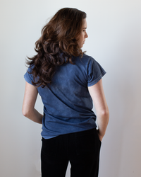 A person with long, wavy brown hair stands facing away, wearing the Free City Canyonkid Circa1972 Supervintage Tee in Moonshrooms Powder and black pants against a plain light background.