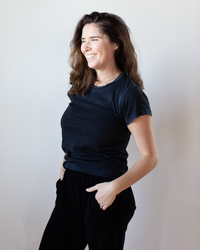 A woman with long brown hair smiles, wearing the Free City Canyonkid Circa1972 Supervintage Tee in Superblack Powder and black pants, with her hands in her pockets, against a plain light background.