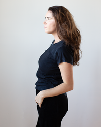 A woman with long brown hair stands in profile against a light background, wearing Free City's Canyonkid Circa1972 Supervintage Tee in Superblack Powder and black pants, one hand in her pocket, showcasing its subtle sunfade dye effect.