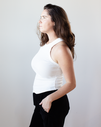 A woman with long brown hair, wearing the NSF Alexa Fitted Baby Rib Tank in Chalk and black pants, stands in profile against a plain light background, looking forward with one hand in her pocket.