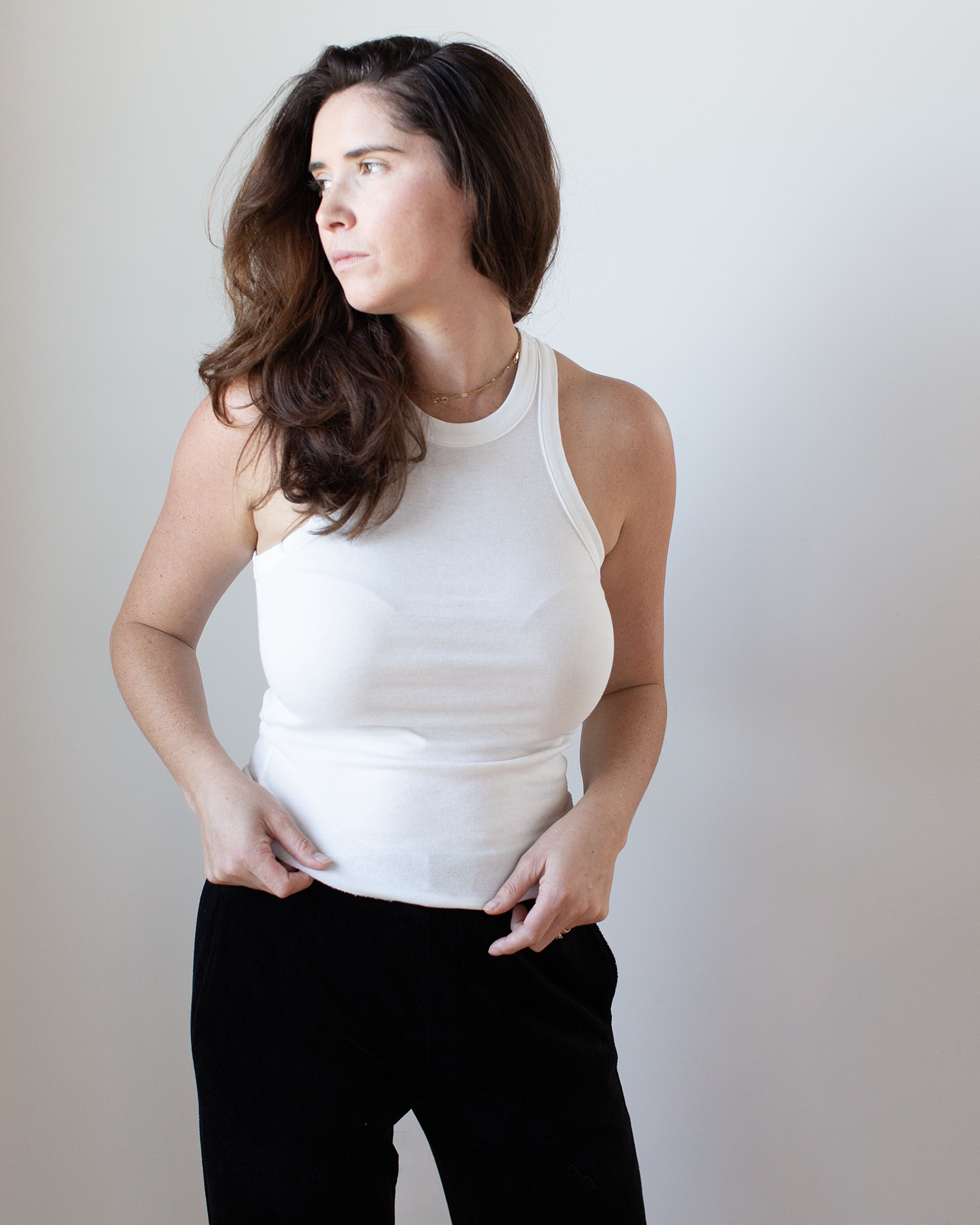 A woman with long brown hair wears the NSF Alexa Fitted Baby Rib Tank in Chalk and black pants, standing against a plain light background, looking to the left.