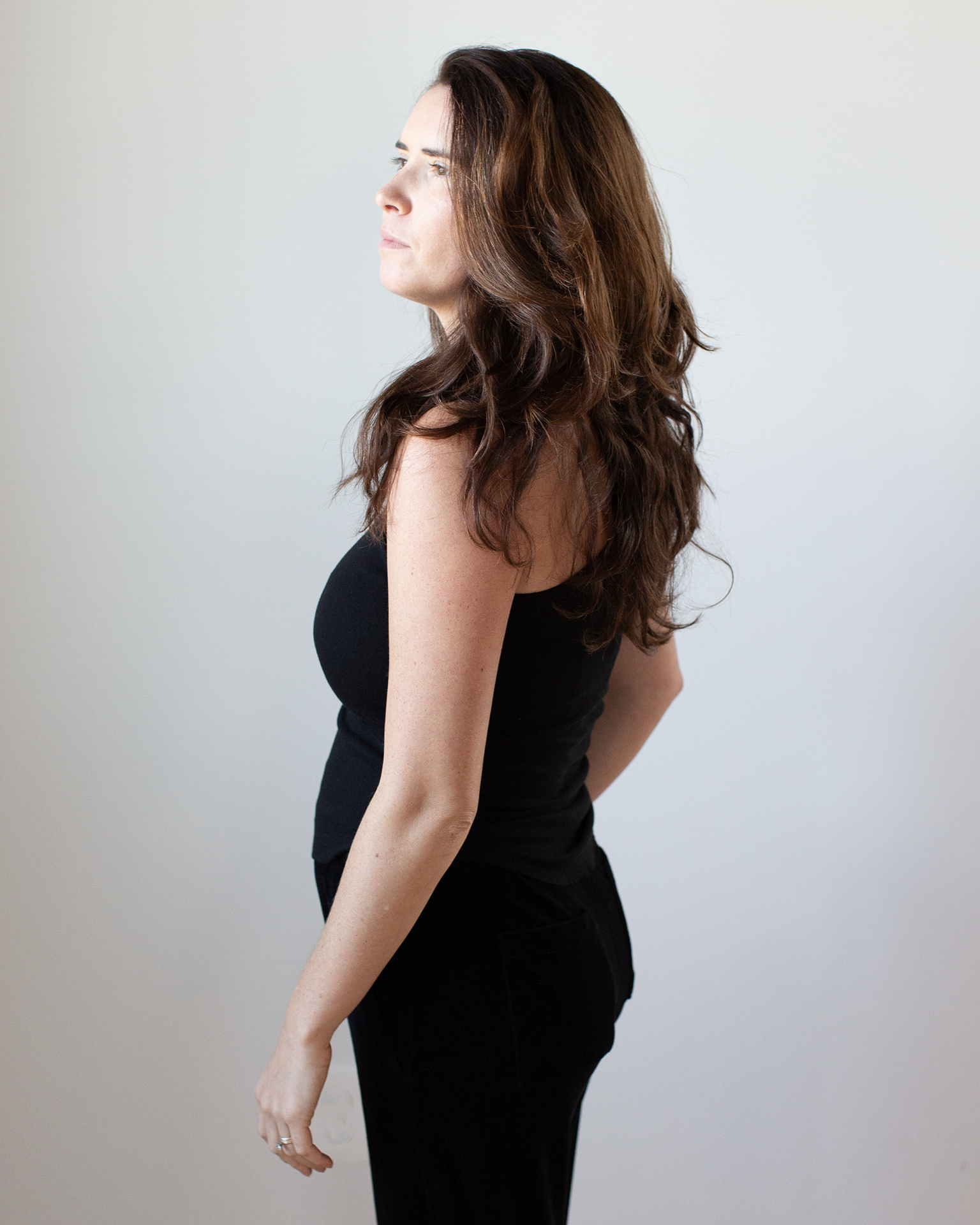 A woman with long brown hair, wearing the NSF Alexa Fitted Baby Rib Tank in Black and black pants, stands in profile against a plain light background, looking slightly upward.