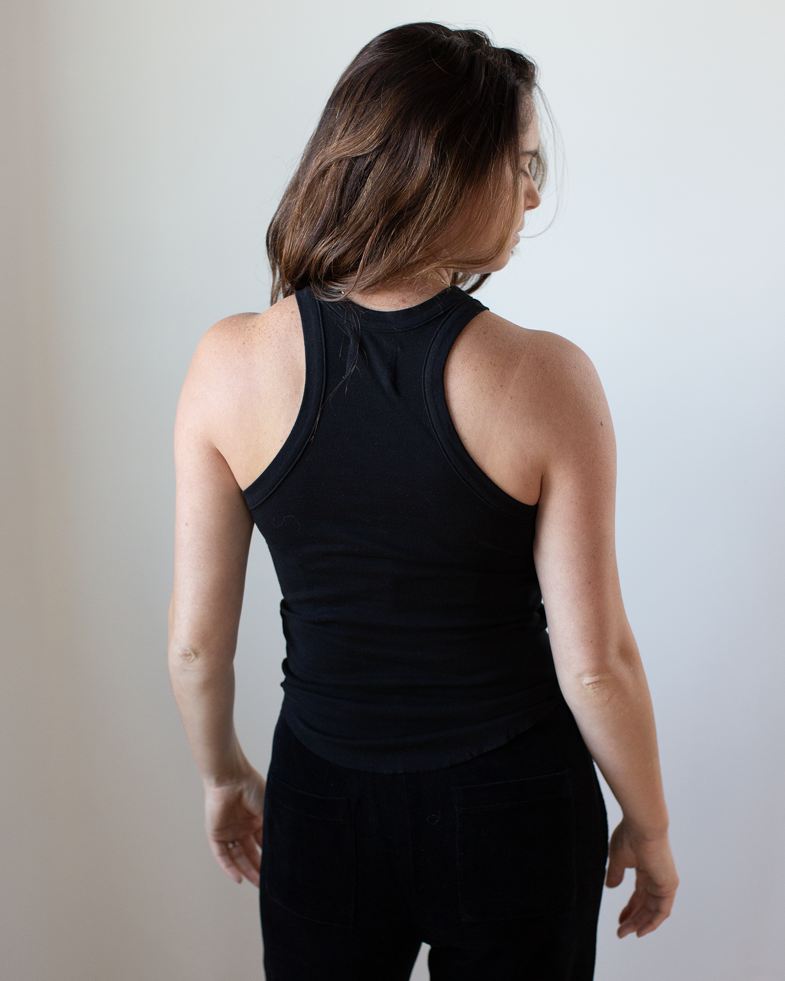 A person with long brown hair stands facing away, wearing the NSF Alexa Fitted Baby Rib Tank in Black and black pants against a plain light background.