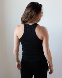 A person with long brown hair stands facing away, wearing the NSF Alexa Fitted Baby Rib Tank in Black and black pants against a plain light background.