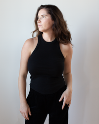 A woman with long brown hair, wearing the NSF Alexa Fitted Baby Rib Tank in Black, stands against a plain light background and looks to her left with a neutral expression.