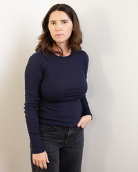 A woman with long brown hair, wearing the Velvet by Graham & Spencer Mauri L/S Crew Neck Top in Navy and black jeans, stands against a plain light wall, looking at the camera with a neutral expression and one hand in her pocket.