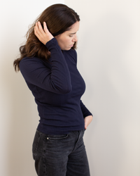 A woman with long brown hair wears the Mauri L/S Crew Neck Top in Navy by Velvet by Graham & Spencer and black jeans, standing against a light background, looking down, touching her hair with one hand and placing the other in her pocket.