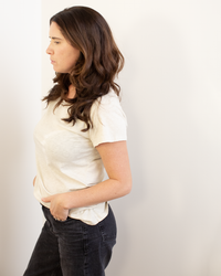 A woman with long brown hair stands in profile against a white wall, wearing the Odelia Scoop Neck Tee in Cookie by Velvet by Graham & Spencer and black jeans, her hands in her pockets.