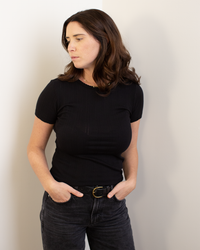 A woman with shoulder-length brown hair wears the Enati S/S Crew Neck Top in Black by Velvet by Graham & Spencer and dark jeans, standing against a white wall with her hands in her pockets, looking to the side with a neutral expression.