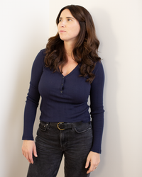 A woman with long brown hair, wearing the Velvet by Graham & Spencer Winona L/S Button Top in Navy and black jeans, stands against a light-colored wall, looking slightly to the side.