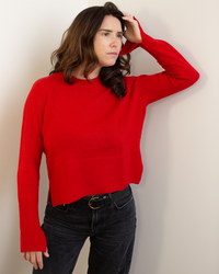 A woman with wavy brown hair wears the Autumn Cashmere Cropped Boxy Crew W/ Slits in Blazer, paired with black jeans, standing against a light wall and looking thoughtfully to the side with one hand touching her head.
