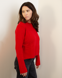 A woman with long brown hair, dressed in Autumn Cashmere’s Cropped Boxy Crew W/ Slits in Blazer and black pants, stands against a pale wall, gazing thoughtfully to the side.