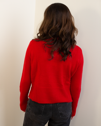 A person with wavy brown hair, wearing the Autumn Cashmere Cropped Boxy Crew W/ Slits in Blazer and dark jeans, stands facing a white wall with their back to the camera.