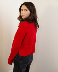 A woman with long brown hair stands sideways, looking over her shoulder at the camera, wearing Autumn Cashmere's Cropped Boxy Crew W/ Slits in Blazer and black jeans against a plain light-colored wall.