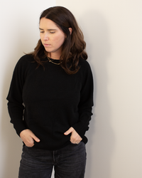 A woman with long brown hair wears the Autumn Cashmere Relaxed Fit Raglan Crew W/ Rib Detail in black and dark jeans, standing against a light-colored wall with her hands in her pockets, looking down.