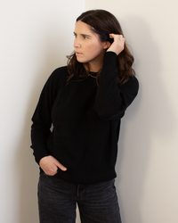 A woman with wavy brown hair models the Autumn Cashmere Relaxed Fit Raglan Crew w/ Rib Detail in Black, paired with dark jeans. She stands against a light wall, looking aside with one hand in her hair and the other in her pocket.