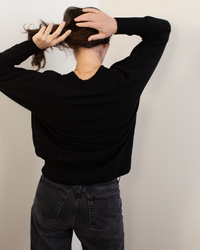 A person with dark hair, wearing Autumn Cashmere's Relaxed Fit Raglan Crew with Rib Detail in Black and dark jeans, stands facing a white wall with their back to the camera, both hands raised as they gather their hair.