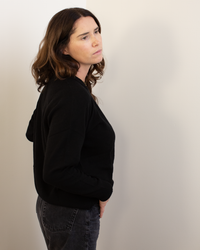 A woman with wavy brown hair, wearing the Autumn Cashmere Relaxed Fit Raglan Crew with Rib Detail in Black and dark jeans, stands sideways against a light wall, gazing thoughtfully into the distance.