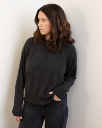 A woman with wavy brown hair wears the Autumn Cashmere Relaxed Fit Raglan Crew W/ Rib Detail in Pepper and black jeans, standing against a light wall with one hand in her pocket, gazing thoughtfully to her left.