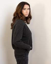 A woman with long brown hair stands sideways, wearing the Autumn Cashmere Relaxed Fit Raglan Crew W/ Rib Detail in Pepper, hands in pockets, against a plain light background, looking seriously at the camera.