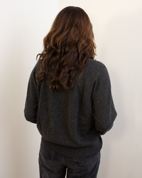 A person with wavy brown hair, seen from behind, wears the Autumn Cashmere Relaxed Fit Raglan Crew W/ Rib Detail in Pepper and dark pants while facing a white wall.